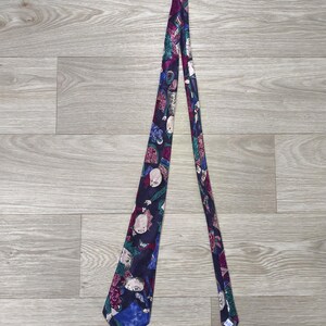 May include: A necktie featuring a dark blue base with a repeating pattern of stylised figures in shades of purple, teal, and white. The design includes abstract shapes and human-like faces distributed across the length of the fabric accessory.