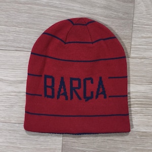 May include: A red knitted beanie hat featuring navy blue horizontal stripes and the word "BARCA" in navy blue lettering. The hat is made of a soft, knitted material and is designed to fit snugly on the head.