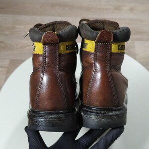 May include: A pair of brown leather work boots with black soles. The boots have yellow accents with the brand name "CATERPILLAR" printed on them. The boots are laced up and appear worn.