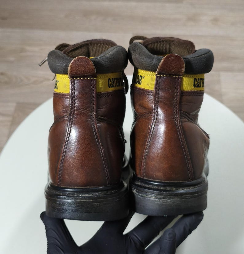 May include: A pair of brown leather work boots with black soles. The boots have yellow accents with the brand name "CATERPILLAR" printed on them. The boots are laced up and appear worn.