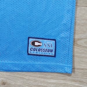 May include: Light blue mesh athletic jersey with a Colosseum Athletics label. The label features the text "XXL" and the brand name. The jersey has a perforated texture.