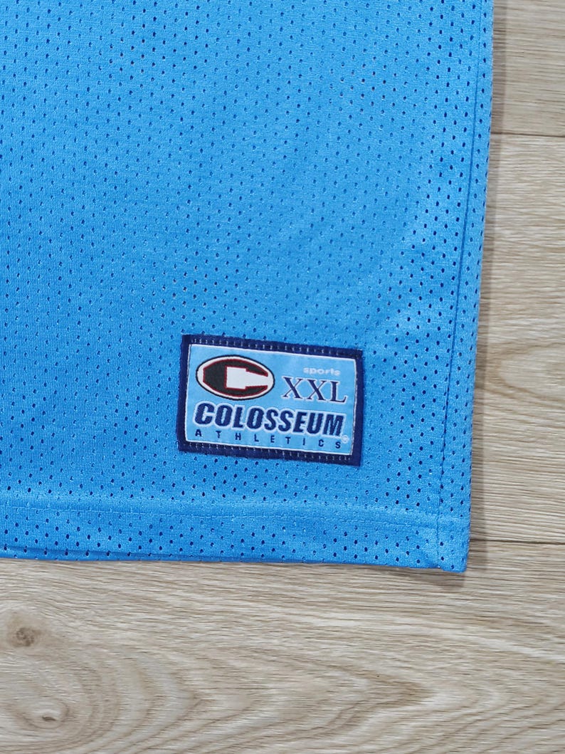 May include: Light blue mesh athletic jersey with a Colosseum Athletics label. The label features the text "XXL" and the brand name. The jersey has a perforated texture.