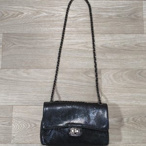 Può includere: Una borsa a tracolla nera con una texture simile alla pelle di serpente, dotata di una patta rettangolare, una chiusura a girello rettangolare color argento e una lunga e sottile tracolla a catena intrecciata con materiale nero.