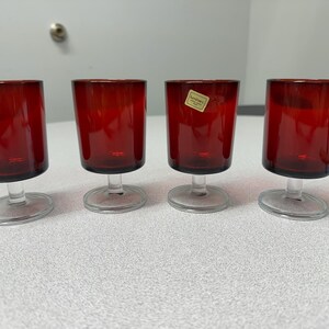 May include: Four ruby red stemmed glasses with clear bases. The glasses have a slightly tapered shape and are evenly spaced on a white surface. The glasses are likely for serving beverages.