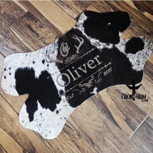 Peut inclure: Un tapis de style peau de vache avec un motif tacheté noir et blanc. Le tapis a une forme unique et porte le nom "Oliver" et l'année "2017" imprimés, ainsi que des éléments décoratifs. Le logo Cross Brim est visible.