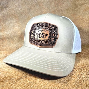 Puede incluir: Gorra de camionero beige y blanca con visera cosida. La gorra presenta un parche de cuero marrón con las palabras "GRAND CHAMPION CASH OUT MARKET STEER" y un gráfico de toro. La gorra tiene una parte trasera de malla.