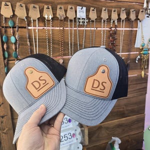 Könnte beinhalten: Zwei graue Trucker-Caps mit schwarzen Mesh-Rücken und Lederaufnähern. Die Aufnäher zeigen die Initialen "DS" in einem rechteckigen Design. Die Caps werden in einem Geschäftsumfeld gezeigt, mit Schmuck im Hintergrund.