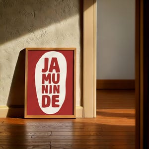 Può includere: Una stampa artistica incorniciata con la scritta "JA MU NIN DE" in bianco su sfondo rosso. La cornice in legno è posta contro un muro strutturato, con la luce del sole che crea ombre sul pavimento in legno.