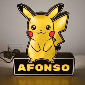 May include: A yellow Pikachu-shaped night light with a black border and base. The base displays the name "AFONSO" in yellow letters. The night light has a black power cord.
