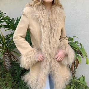 May include: A tan suede coat with a shaggy, light brown fur collar, cuffs, and trim. The coat has long sleeves and a tailored fit. The fur trim adds a luxurious texture to the outerwear. The coat is a vintage style.