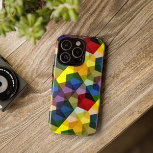 Könnte beinhalten: Eine Smartphone-Hülle mit einem farbenfrohen geometrischen Muster. Die Hülle zeigt ein Mosaikmuster in Gelb-, Rot-, Blau-, Lila- und Grüntönen. Das Telefon liegt auf einer Holzoberfläche, im Hintergrund sind eine Kamera und eine Pflanze zu sehen.