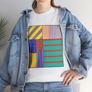 Könnte beinhalten: Weißes T-Shirt mit einem farbenfrohen geometrischen Design aus Streifen, Punkten und verschiedenen Formen in Gelb-, Rosa-, Blau- und Grüntönen. Das Shirt wird mit einer blauen Jeansjacke getragen.