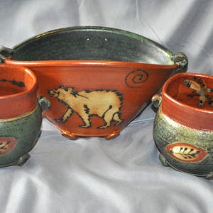 May include: A ceramic set consisting of one large bowl and two smaller matching cups. The pieces feature a two-tone glaze with a dark green base and a reddish-orange rim. The bowl displays a bear illustration, while the cups feature leaf motifs.