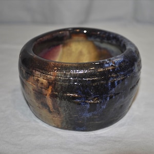 May include: A small, round ceramic bowl with a thick, rounded rim. The exterior features a dark, glossy glaze with blended tones of black, deep blue, and streaks of warm brown and tan, creating a textured, multi-colored appearance.