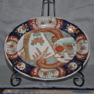 May include: An oval ceramic plate with a detailed Asian-inspired design. The plate features a white center with bamboo and floral motifs, framed by a blue border with orange and gold accents. It is displayed on a black metal stand.