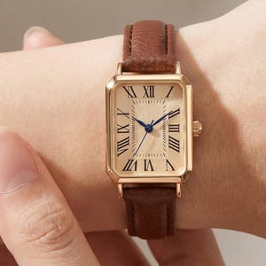 Puede incluir: Un reloj de pulsera rectangular con caja en tono dorado y esfera color champán texturizada con números romanos negros. El reloj tiene manecillas azules delgadas y está sujeto a una correa de cuero marrón con textura granulada visible.