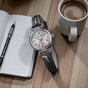 May include: A silver OLEVS quartz watch with a black leather band, Roman numeral hour markers, and a crown detail. The watch rests on a wooden surface next to a notebook, pen, and a cup of coffee.