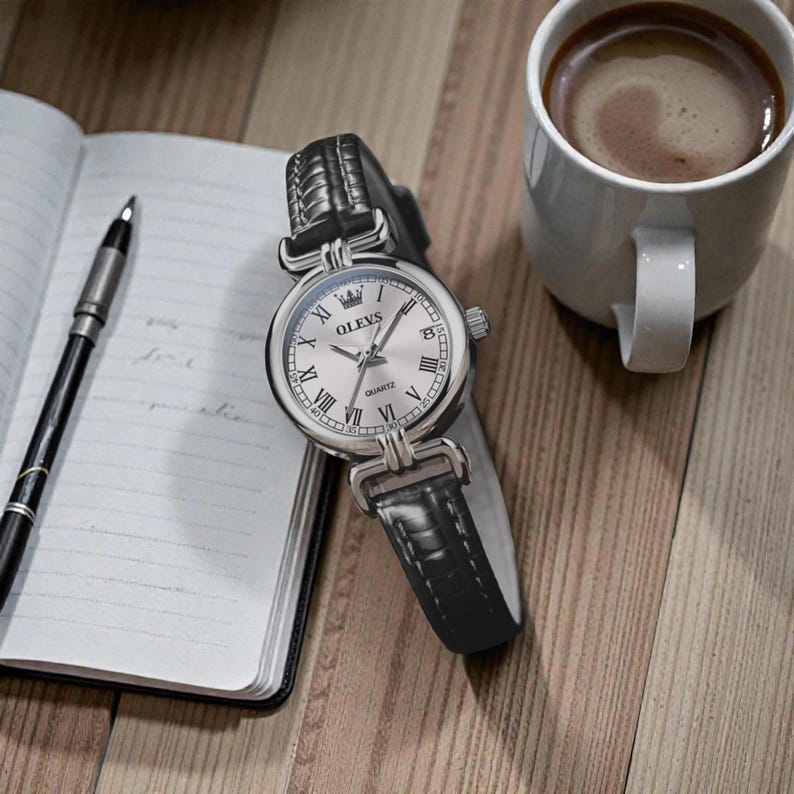 May include: A silver OLEVS quartz watch with a black leather band, Roman numeral hour markers, and a crown detail. The watch rests on a wooden surface next to a notebook, pen, and a cup of coffee.