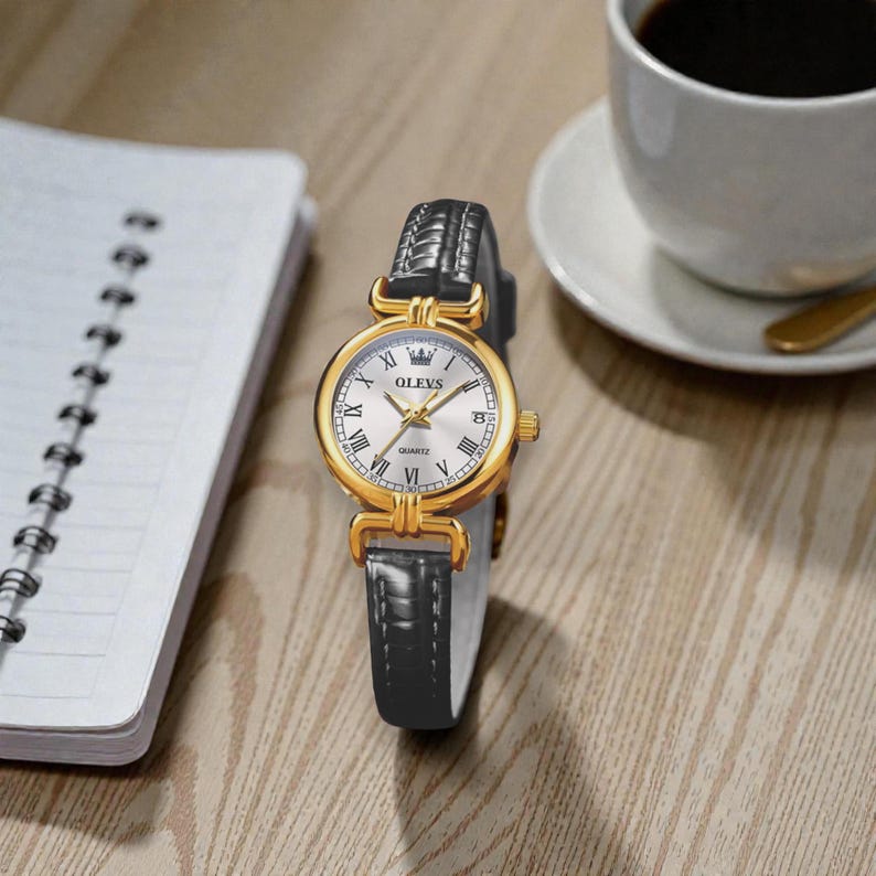 May include: Gold-tone OLEVS quartz watch with a white face, Roman numeral hour markers, and a date display. The watch has a black leather band and is displayed on a wooden surface. A cup of coffee and a notebook are also visible.