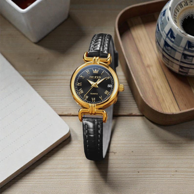 May include: A gold-toned OLEVS quartz watch with a black dial and Roman numeral hour markers. The watch features a date display and a black leather strap. The watch is displayed on a wooden surface.