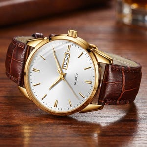 May include: A gold-tone quartz wristwatch featuring a round white dial with gold-tone hour markers and hands. The watch includes a day and date display window at the three o'clock position and is fitted with a dark brown crocodile-embossed leather strap.