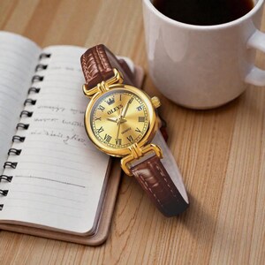 May include: A gold-toned OLEVS quartz watch with a brown leather band rests on a notebook. The watch features Roman numeral hour markers and a date display. A white coffee mug is in the background.