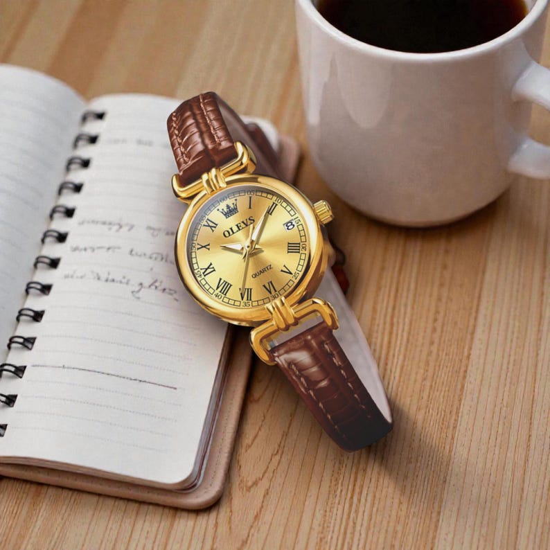 May include: A gold-toned OLEVS quartz watch with a brown leather band rests on a notebook. The watch features Roman numeral hour markers and a date display. A white coffee mug is in the background.