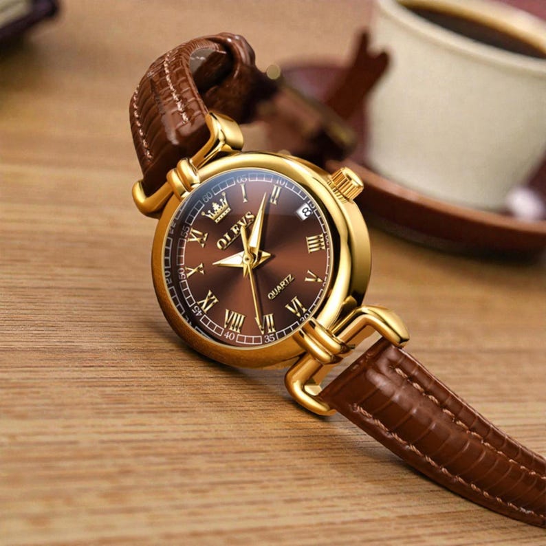 May include: A gold-toned quartz watch with a brown face and a brown leather band. The watch features Roman numeral hour markers and a date display. The watch is set against a wooden surface, with a cup of coffee in the background.