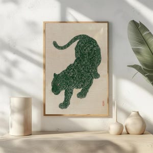 May include: A framed art print featuring a green jaguar illustration. The jaguar is depicted in a walking pose against a cream-colored background. The artwork has a vintage aesthetic, with a gold frame and a small red stamp in the lower right corner.
