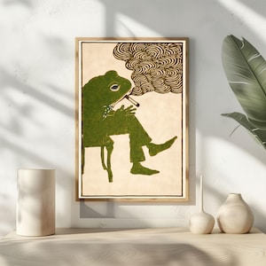 May include: A framed art print featuring a green frog in a suit, smoking a cigarette while seated. The frog is depicted with a blue bow tie and is set against a cream-colored background. The artwork has a vintage, illustrative style.