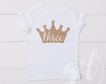 May include: White t-shirt with a gold glitter crown design and the word "three" in a script font. The shirt is a short-sleeved style, perfect for a child's third birthday celebration.