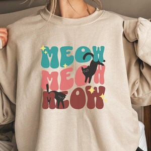 May include: Beige sweatshirt with the words "MEOW MEOW MEOW" in teal, pink, and maroon, with black cat illustrations. The sweatshirt has a crew neck and long sleeves.