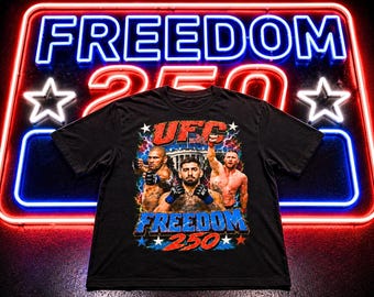 Puede incluir: Camiseta negra de la UFC con un gráfico que presenta a tres luchadores, el logotipo de la UFC y el texto "FREEDOM 250". El diseño incorpora colores rojo, blanco y azul con acentos de rayos. El fondo es un letrero de neón que dice "FREEDOM 250".