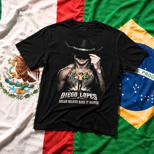 Pode incluir: Uma t-shirt preta de manga curta com uma impressão gráfica de uma pessoa a usar um chapéu de cowboy, com o texto "DIEGO LOPES" e "DREAM BELIEVE MAKE IT HAPPEN" impresso abaixo da imagem. A t-shirt é exibida sobre um fundo de bandeiras.