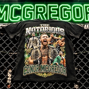 May include: A black t-shirt featuring a graphic print of Conor McGregor. The design includes the text THE NOTORIOUS and CONOR MCGREGOR, along with multiple images of the athlete, including one wearing a championship belt and one with an Irish flag.
