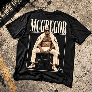 May include: A black short-sleeve t-shirt featuring a graphic print of a person sitting on a stool, wearing a white coat and trousers, with the text MCGREGOR in large white letters above the image and the initials CM in the bottom right corner.