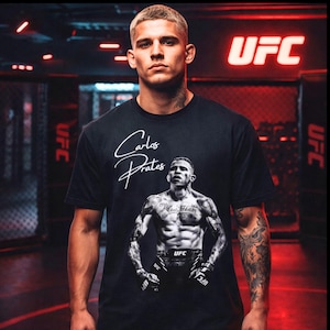 May include: Black t-shirt featuring a portrait of Carlos Prates, with his name in white script. The UFC logo is visible on the shirt and in the background. The image is of a man wearing the shirt.