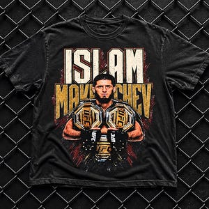 Könnte beinhalten: Ein schwarzes Kurzarm-T-Shirt mit einem Grafikdruck von Islam Makhachev, der zwei goldene UFC-Meisterschaftsgürtel hält. Der Text ISLAM MAKHACHEV ist in großen, abgenutzten weißen und gelben Buchstaben über dem Bild abgedruckt.