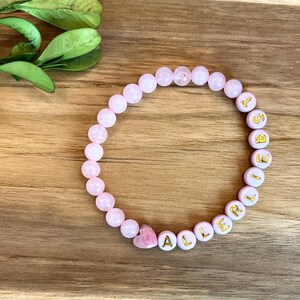 May include: A pink beaded bracelet with the word "ALERT" spelled out in gold letters. The bracelet includes a pink heart-shaped bead. The bracelet is on a wooden surface with green leaves in the background.