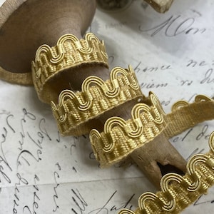 May include: Gold trim with a scalloped edge. The trim is wound around a wooden spool and is laying on a piece of paper with handwritten text.