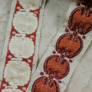 Pode incluir: Duas peças de guarnição vintage com fundo branco e bordado marrom e laranja. Uma guarnição apresenta um padrão repetido de círculos e a outra apresenta um padrão repetido de folhas estilizadas.