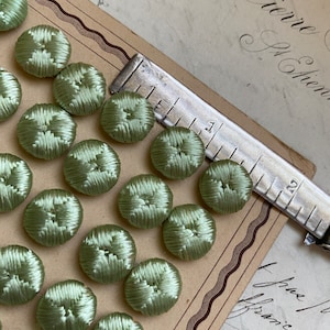 Pode incluir: Uma coleção de 18 botões cobertos de tecido verde-sálvia com um design central em relevo. Os botões estão dispostos em filas em um cartão marrom com uma régua de prata.