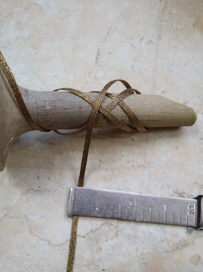 May include: A gold-colored, woven ribbon is wrapped around a wooden form. A metal ruler is positioned next to the ribbon, showing measurements in inches. The ribbon appears to be approximately 1/8 inch wide.