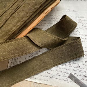 May include: A close-up of a green patterned trim with a geometric design. The trim is folded and stacked on top of a piece of paper with handwritten text.