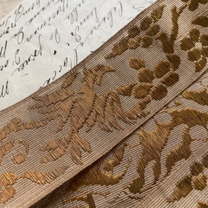 Puede incluir: Primer plano de una cinta beige con un patrón floral metálico dorado. La cinta está tejida con una superficie texturizada y tiene un brillo sutil.