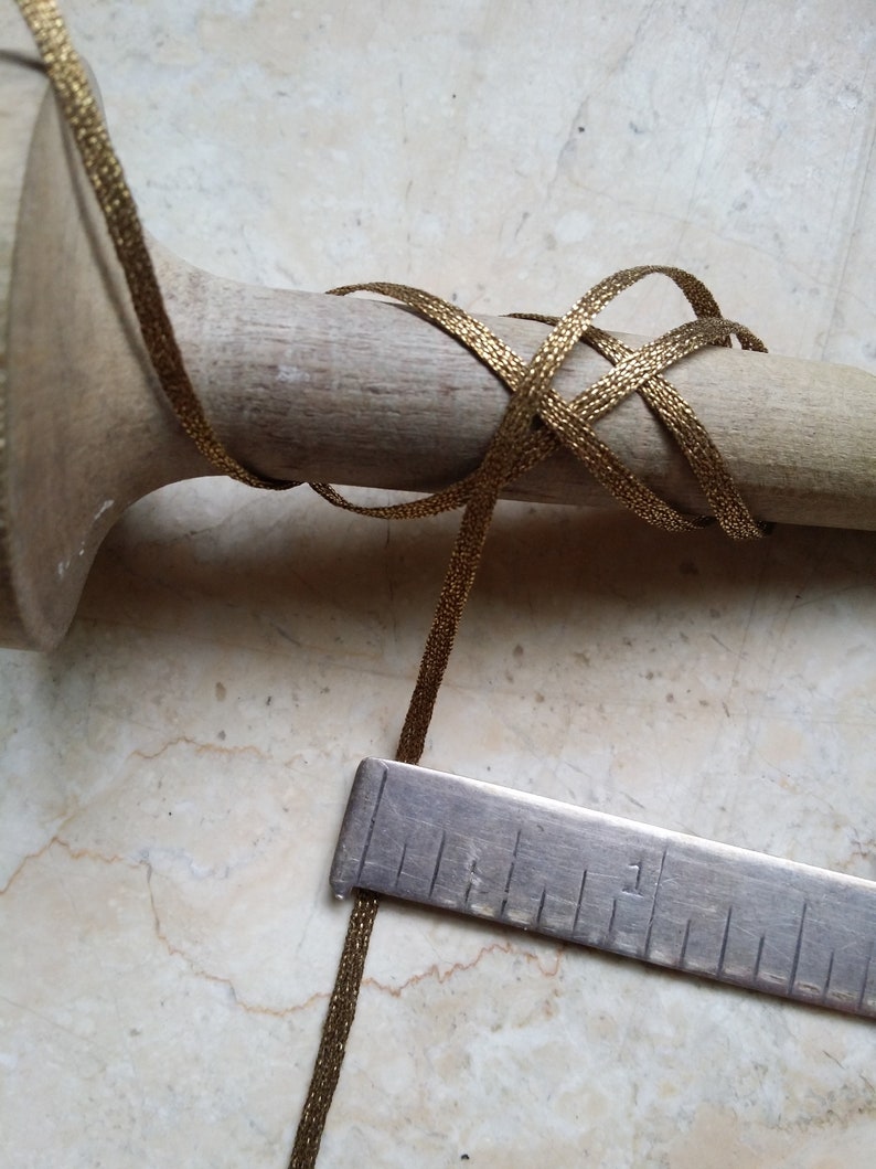 May include: Close-up of a gold metallic ribbon wrapped around a wooden dowel. A metal ruler is placed next to the ribbon. The ribbon has a woven texture and is a warm gold color. The dowel is light brown.