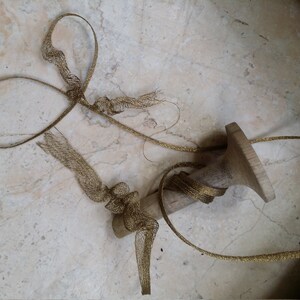 May include: A wooden candlestick with a gold mesh ribbon wrapped around it. The ribbon is knotted in several places and extends across a light-colored, marbled surface. The candlestick has a wide base and a narrow top.