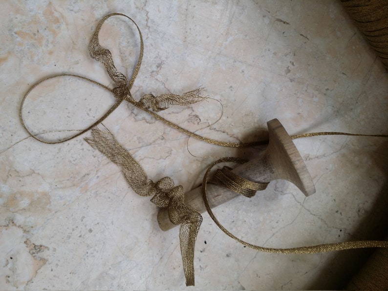 May include: A wooden candlestick with a gold mesh ribbon wrapped around it. The ribbon is knotted in several places and extends across a light-colored, marbled surface. The candlestick has a wide base and a narrow top.