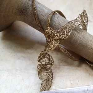 May include: Close-up of a gold-colored, open-weave ribbon tied in a bow. The ribbon is draped over a wooden dowel. A metal ruler is in the foreground, showing measurements in inches.
