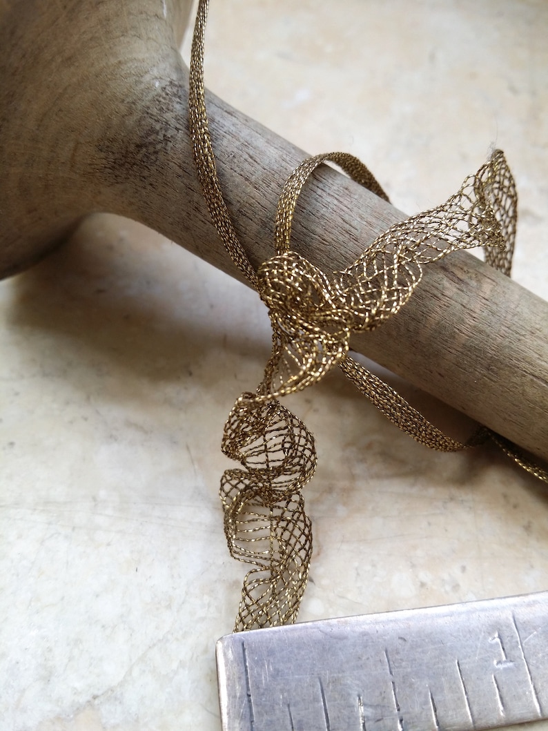 May include: Close-up of a gold-colored, open-weave ribbon tied in a bow. The ribbon is draped over a wooden dowel. A metal ruler is in the foreground, showing measurements in inches.